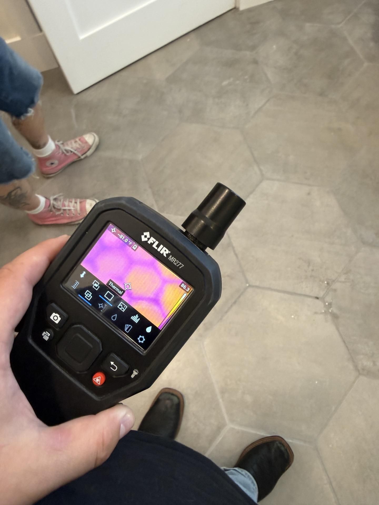 Handheld thermal camera showing a colorful heat map on a tiled floor beside people’s feet