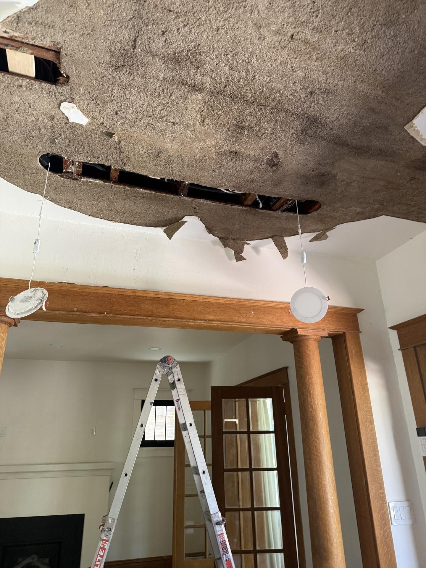 Partially collapsed ceiling with exposed insulation in a room, ladder below and a wooden beam over a doorway