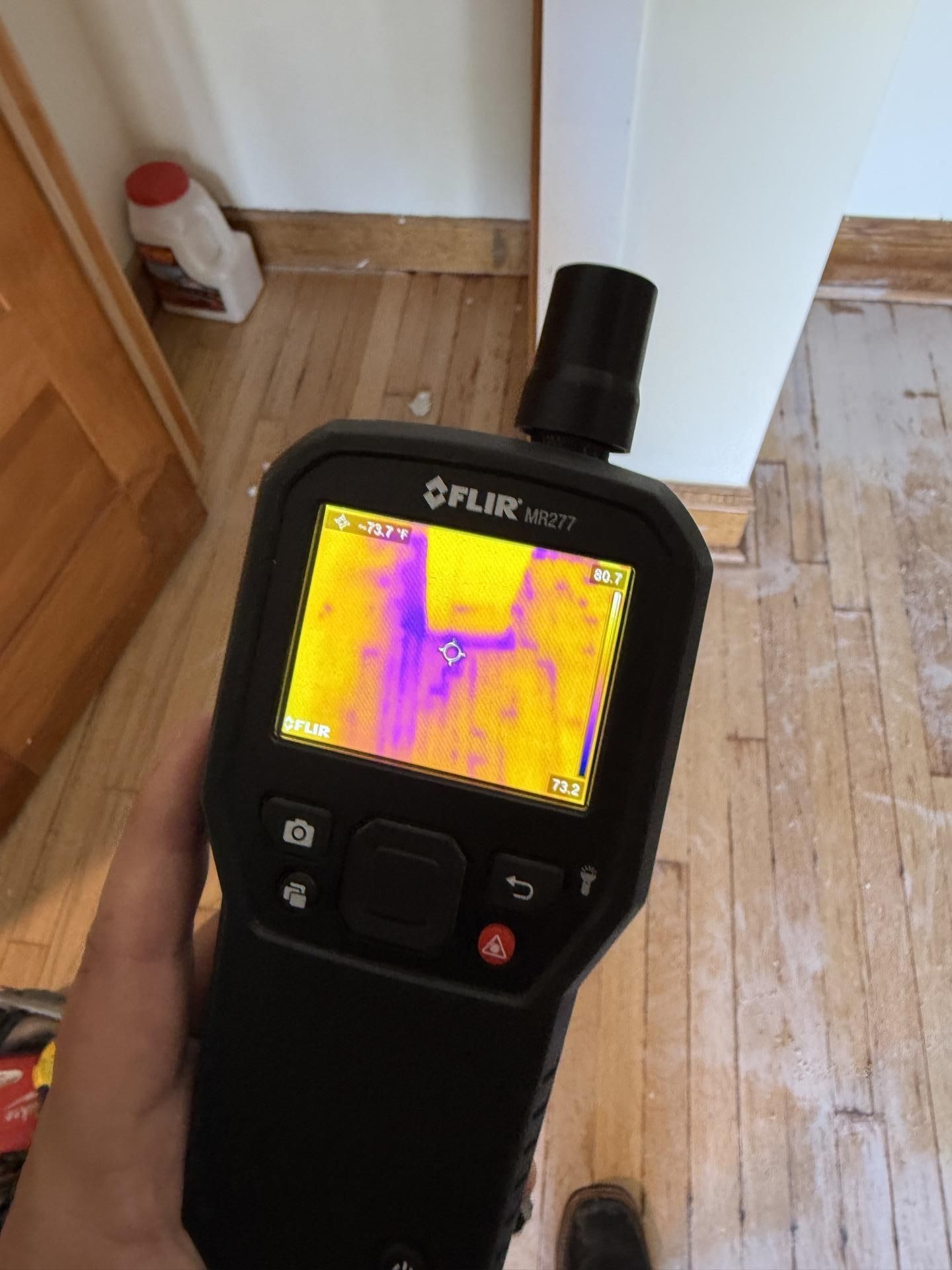 Handheld thermal camera showing a colorful heat map in a room with wooden floor and a door