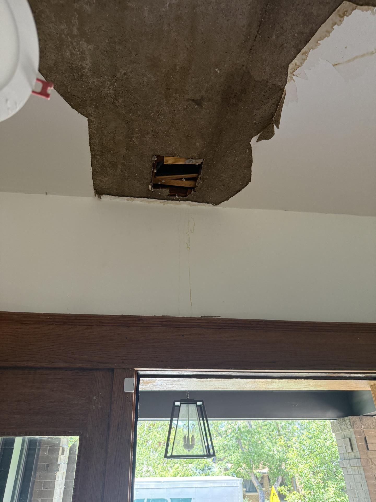 Damaged ceiling above a doorway with exposed patch and hanging electrical box, daylight visible through glass door