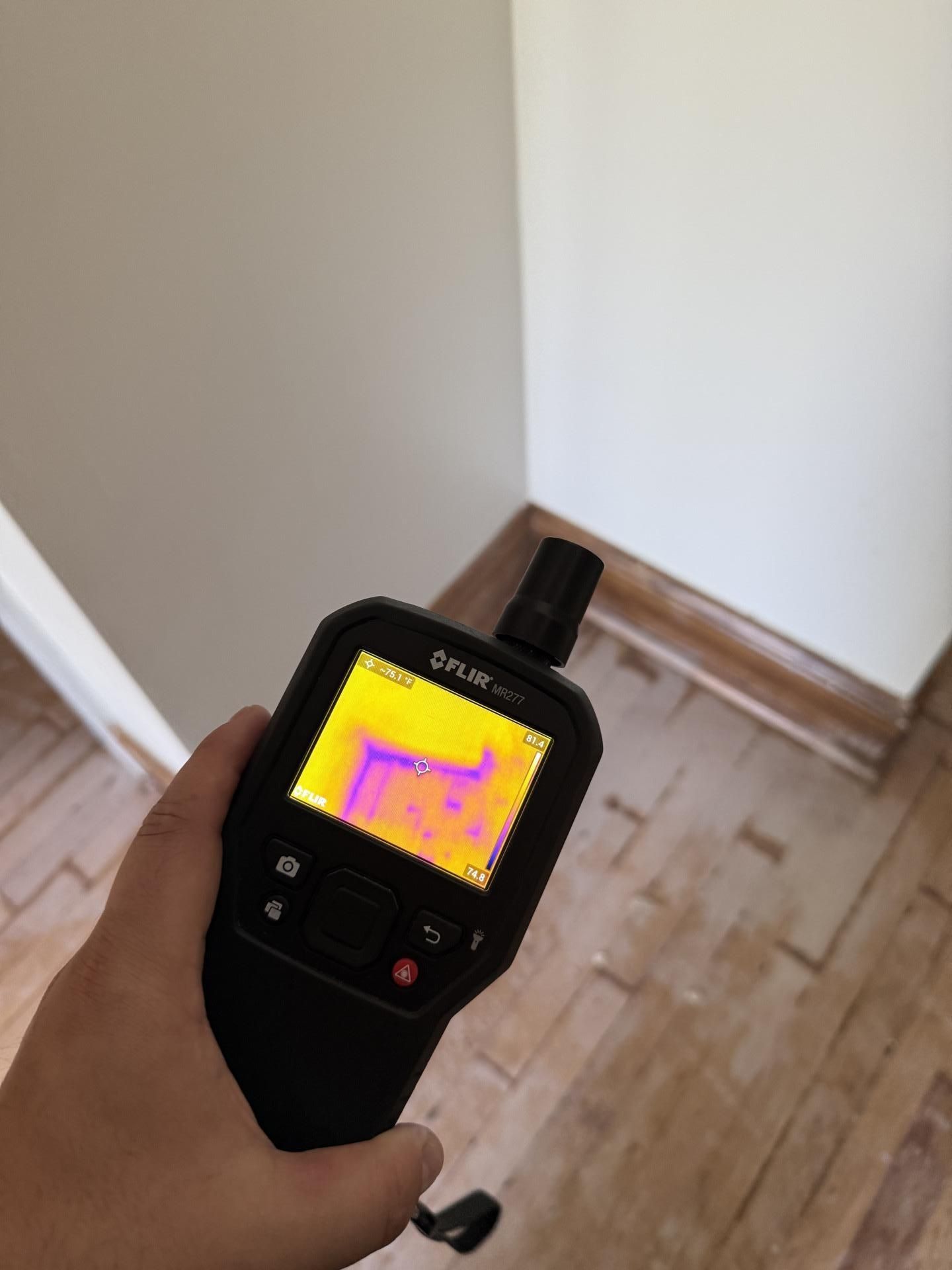 Handheld thermal camera aimed at a room corner, showing a heat map on its screen.