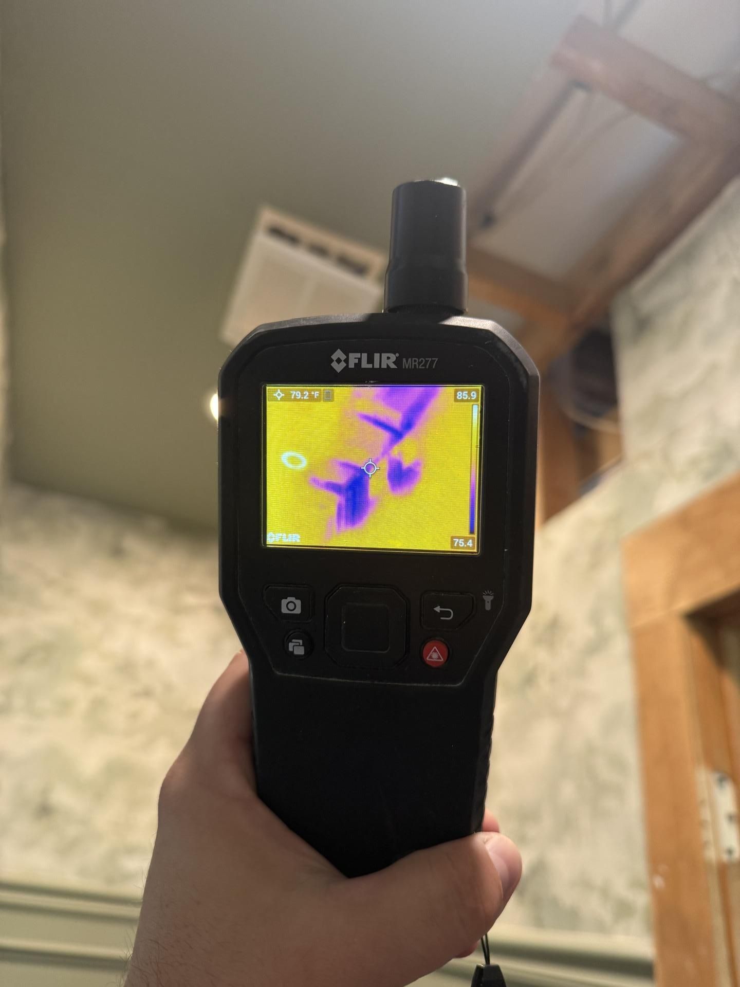 Handheld FLIR thermal camera showing a purple and yellow heat map in a room.