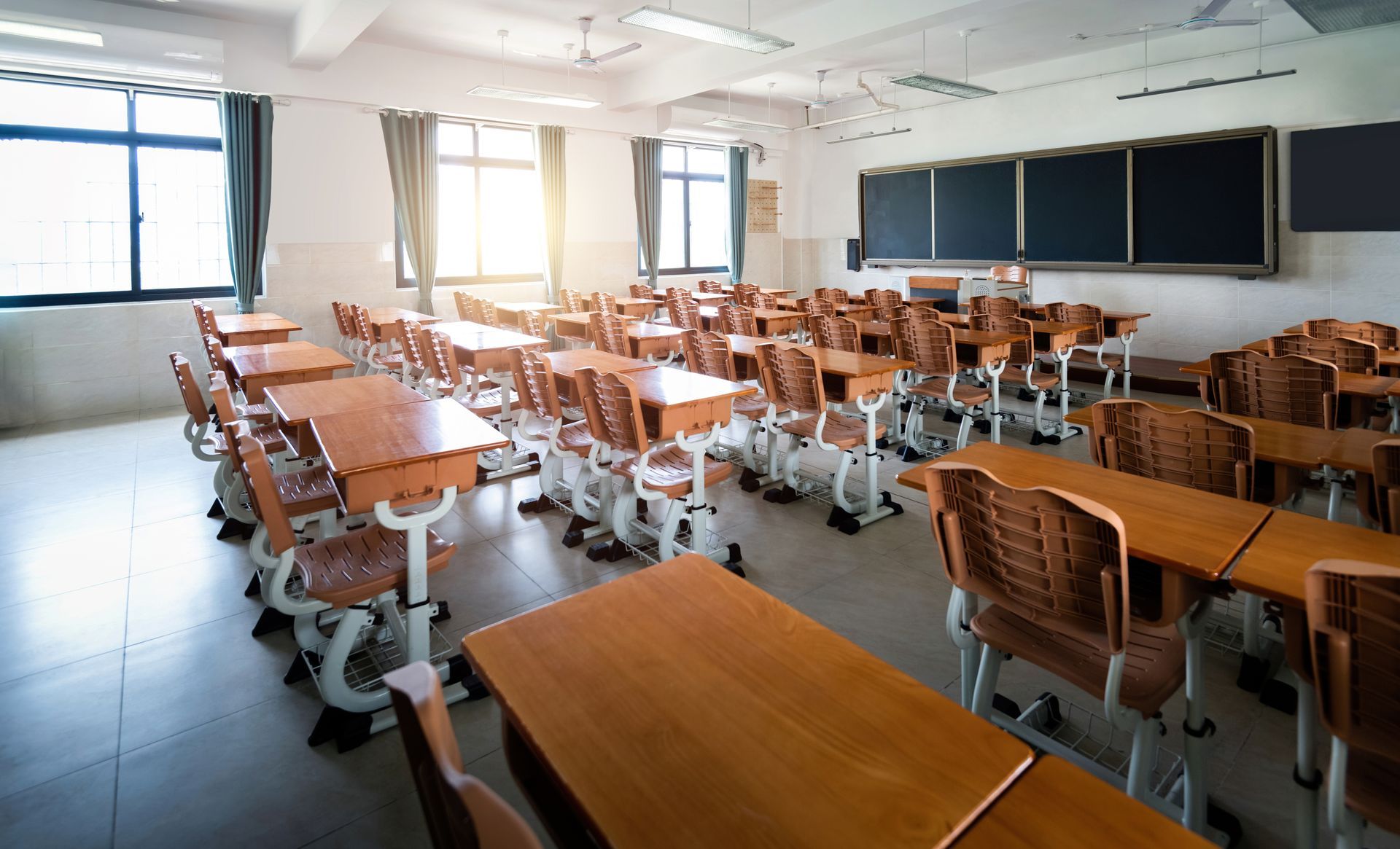 Empty classroom with rows of wooden desks, large windows, and a chalkboard at the front