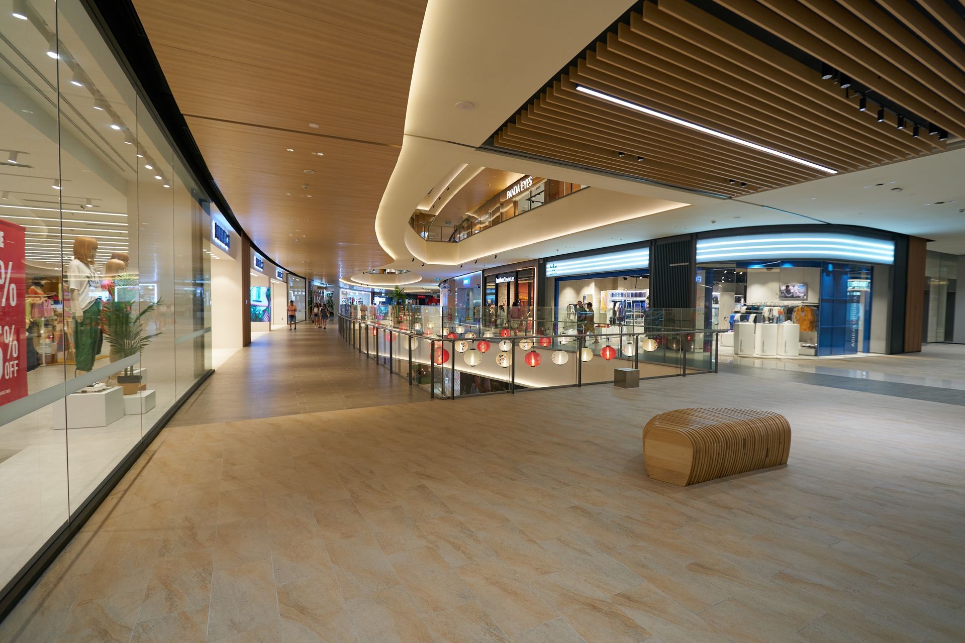 Empty indoor shopping mall corridor with storefronts, glass railings, and a woven bench on the floor