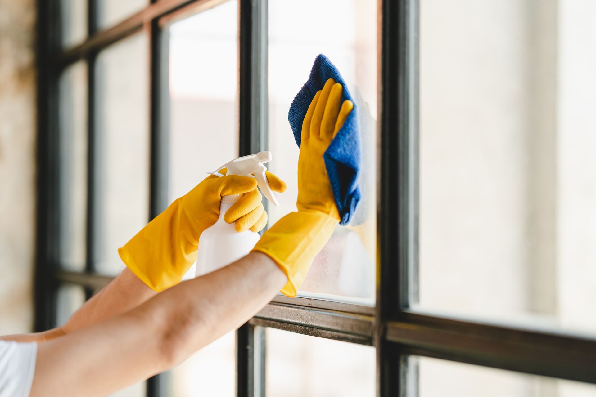 Gloved hands cleaning a window with a spray bottle and blue cloth