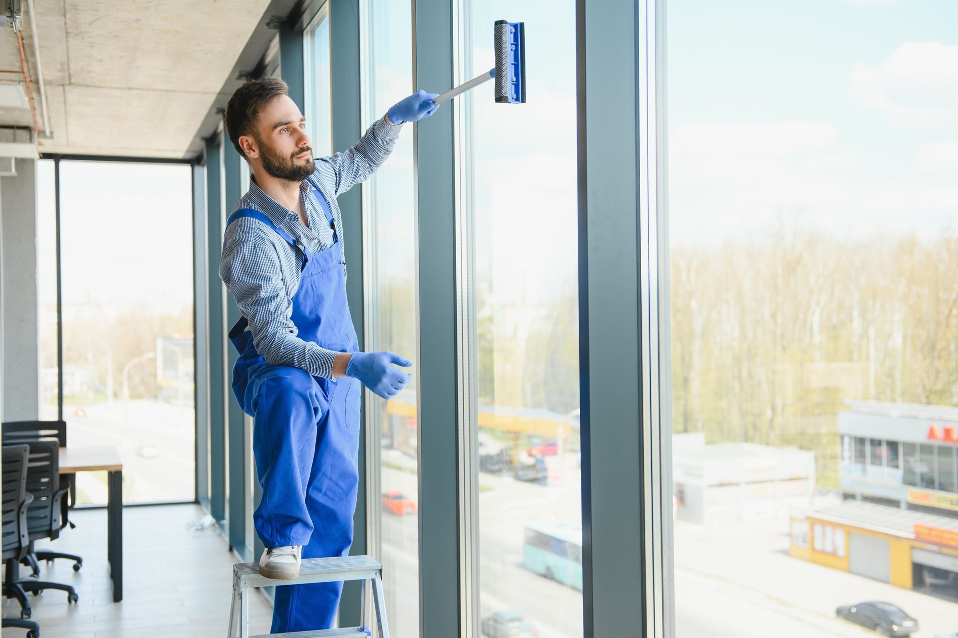 Window cleaner on a step ladder cleaning large glass windows in a bright office interior