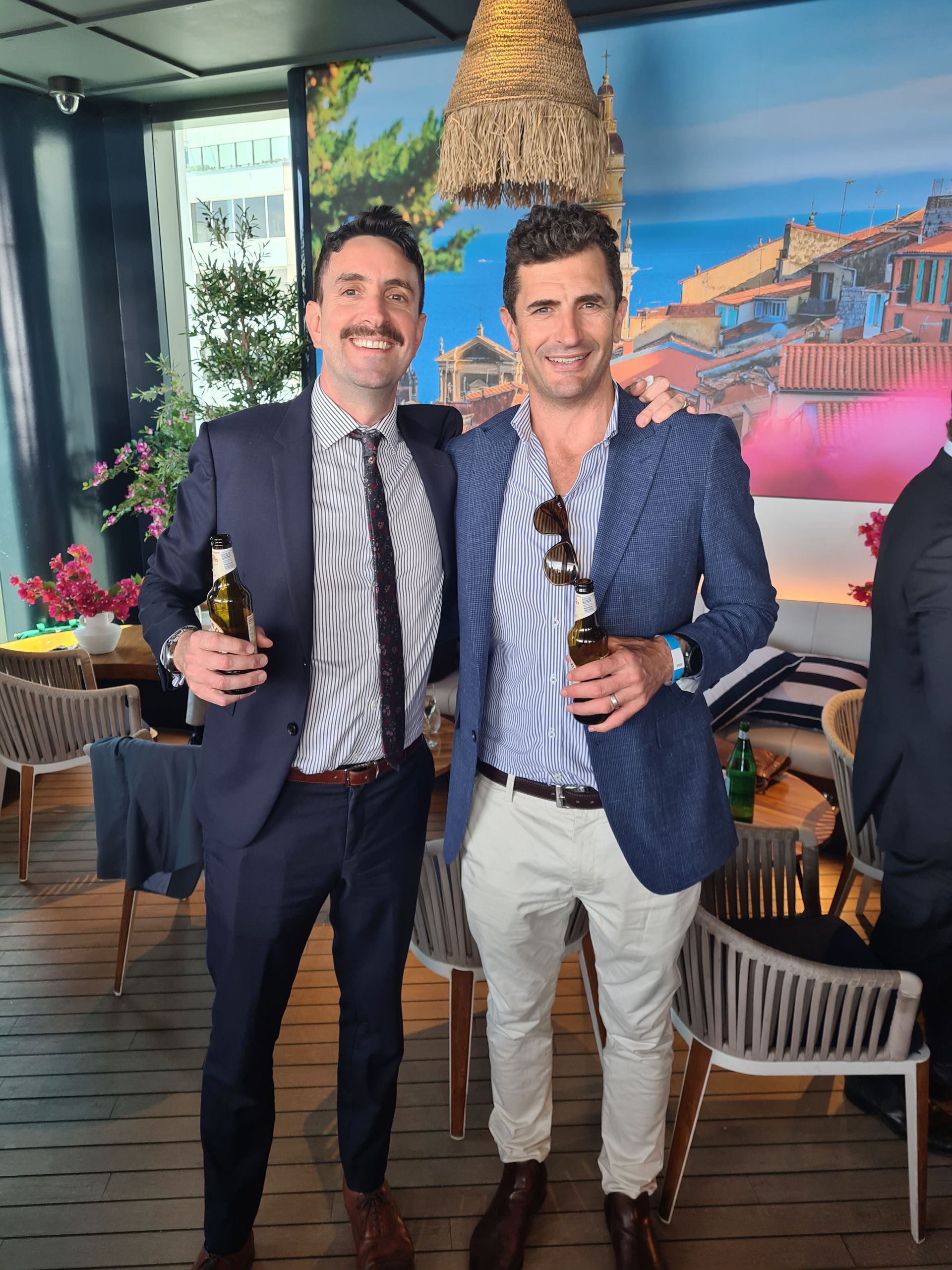 Two men in blazers holding drinks and smiling in a restaurant with a lit Eiffel Tower backdrop