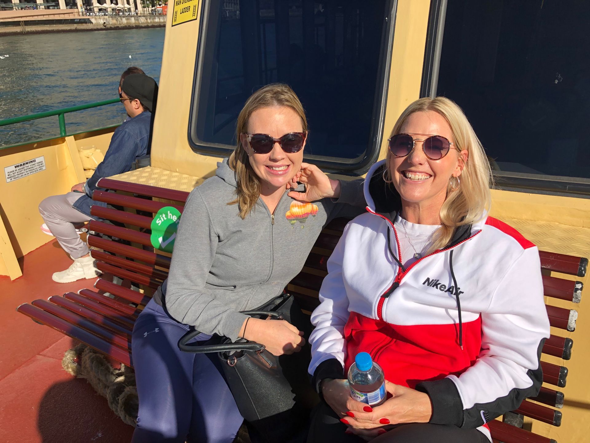 Two women smiling on a ferry bench, one in gray and one in red and white, with water behind them.