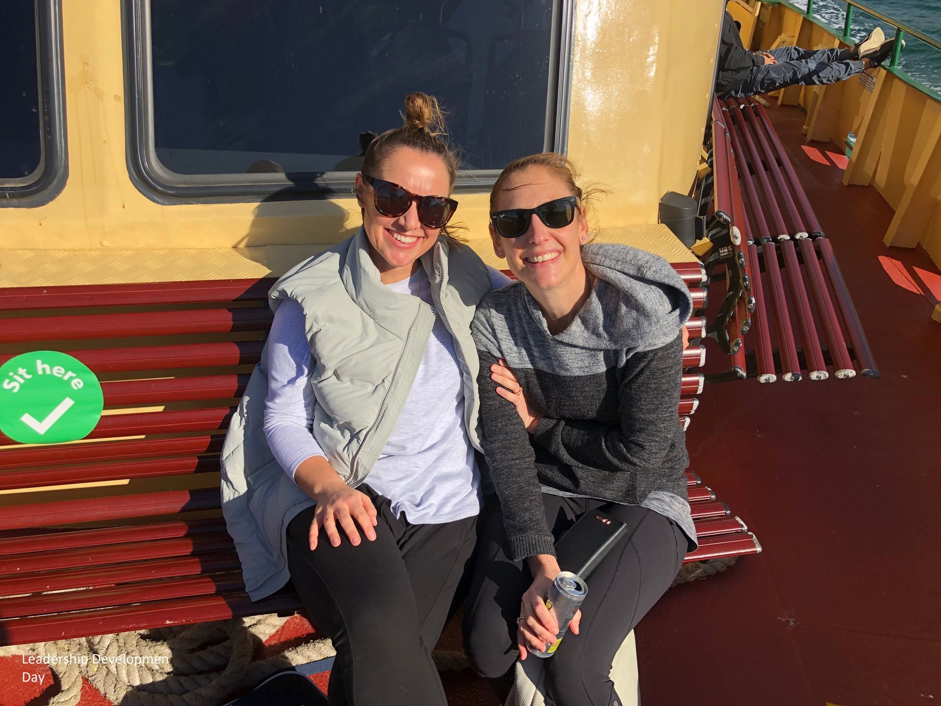 Two people sitting together on a red bench outside a yellow tram, smiling in sunglasses.