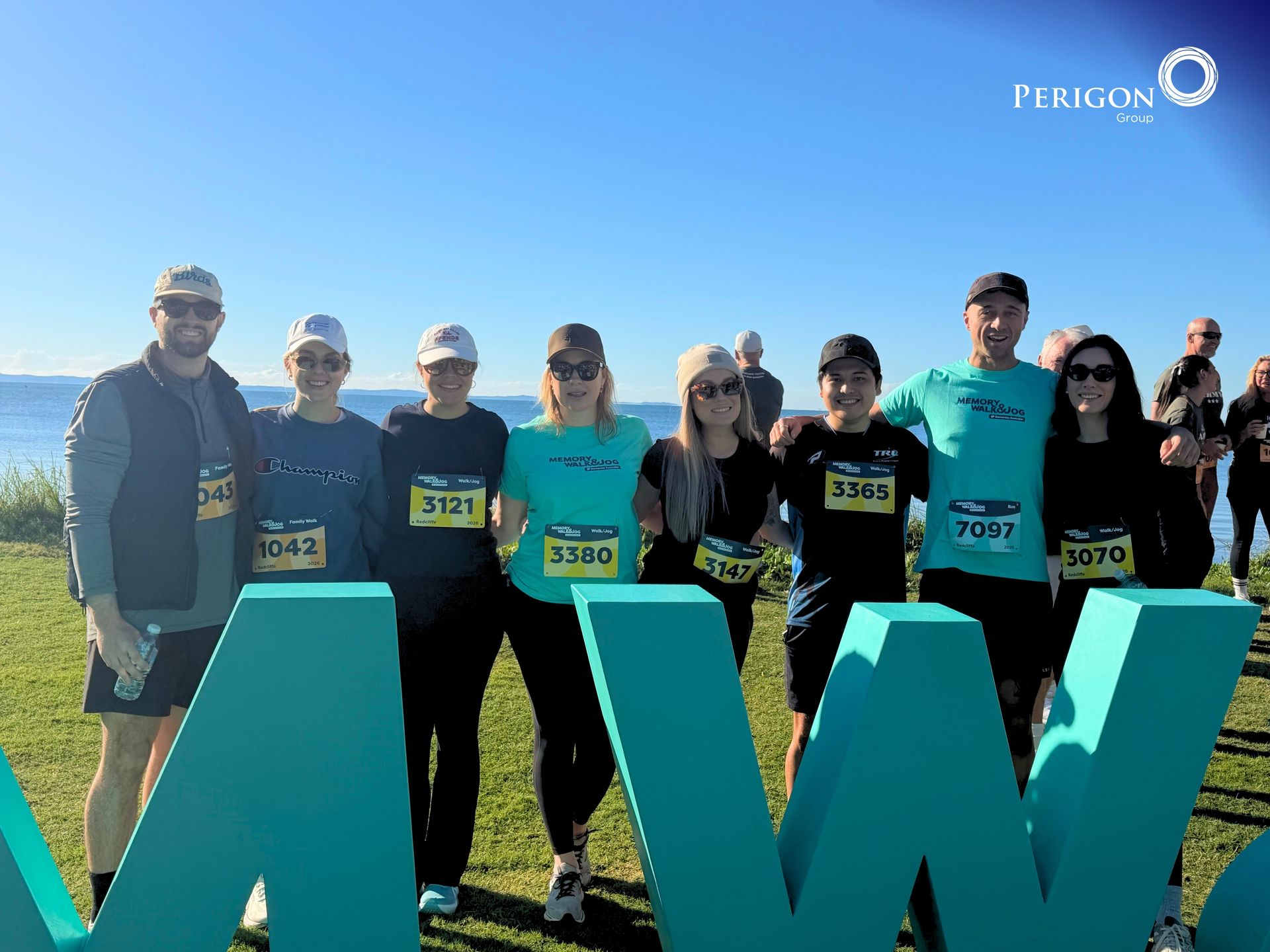 Group of runners posing behind large teal letters at a sunny outdoor event.