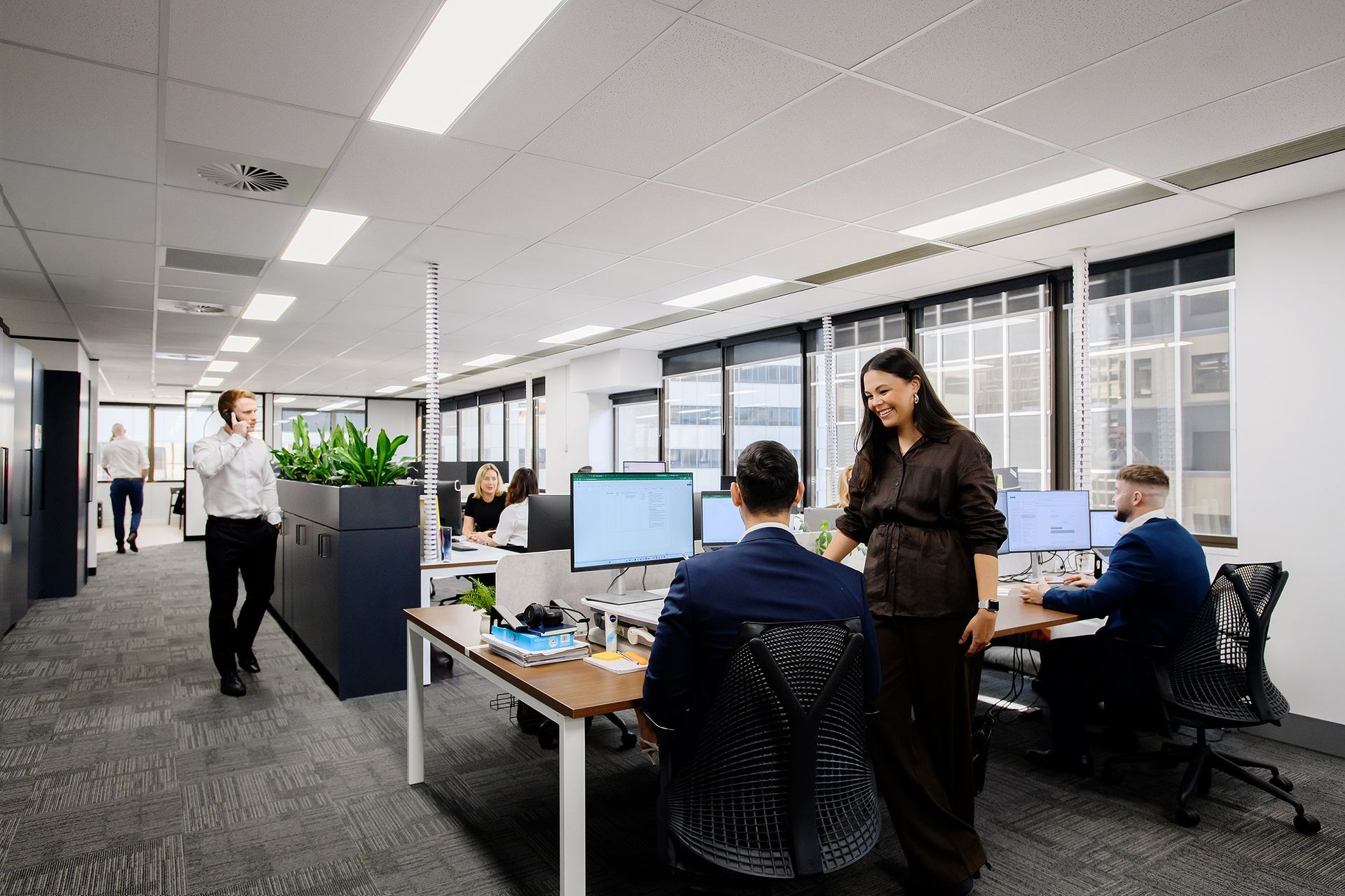 Open-plan office with several coworkers at desks, computers, and a standing discussion near a large window.