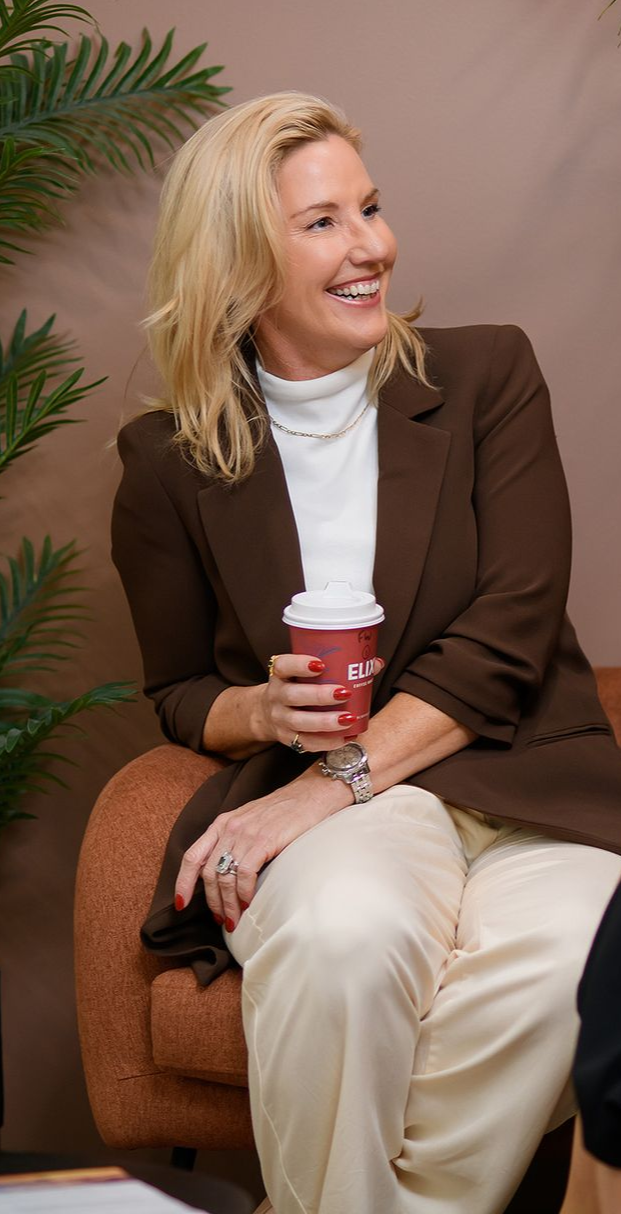 Woman in brown blazer and cream pants holding a coffee cup, smiling while seated indoors.