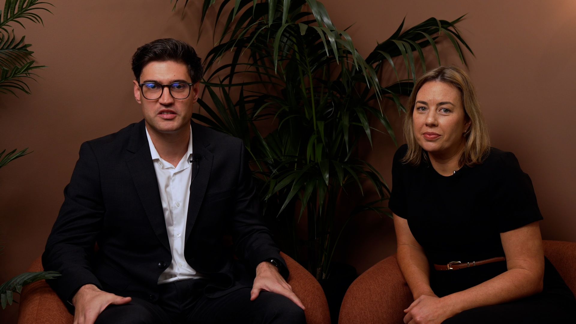 Two people seated indoors against a brown wall with a large leafy plant behind them.