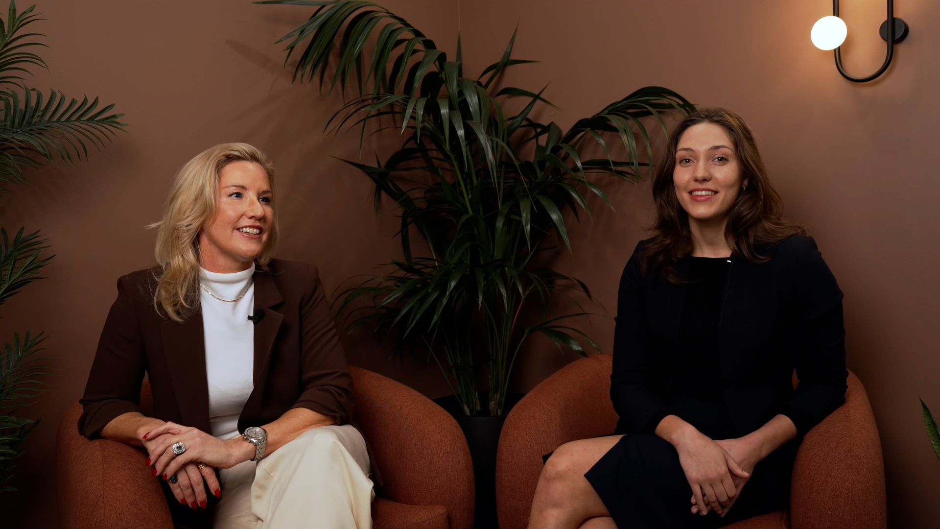 Two women seated in armchairs, smiling in a warmly lit indoor lounge with plants and wall sconces.