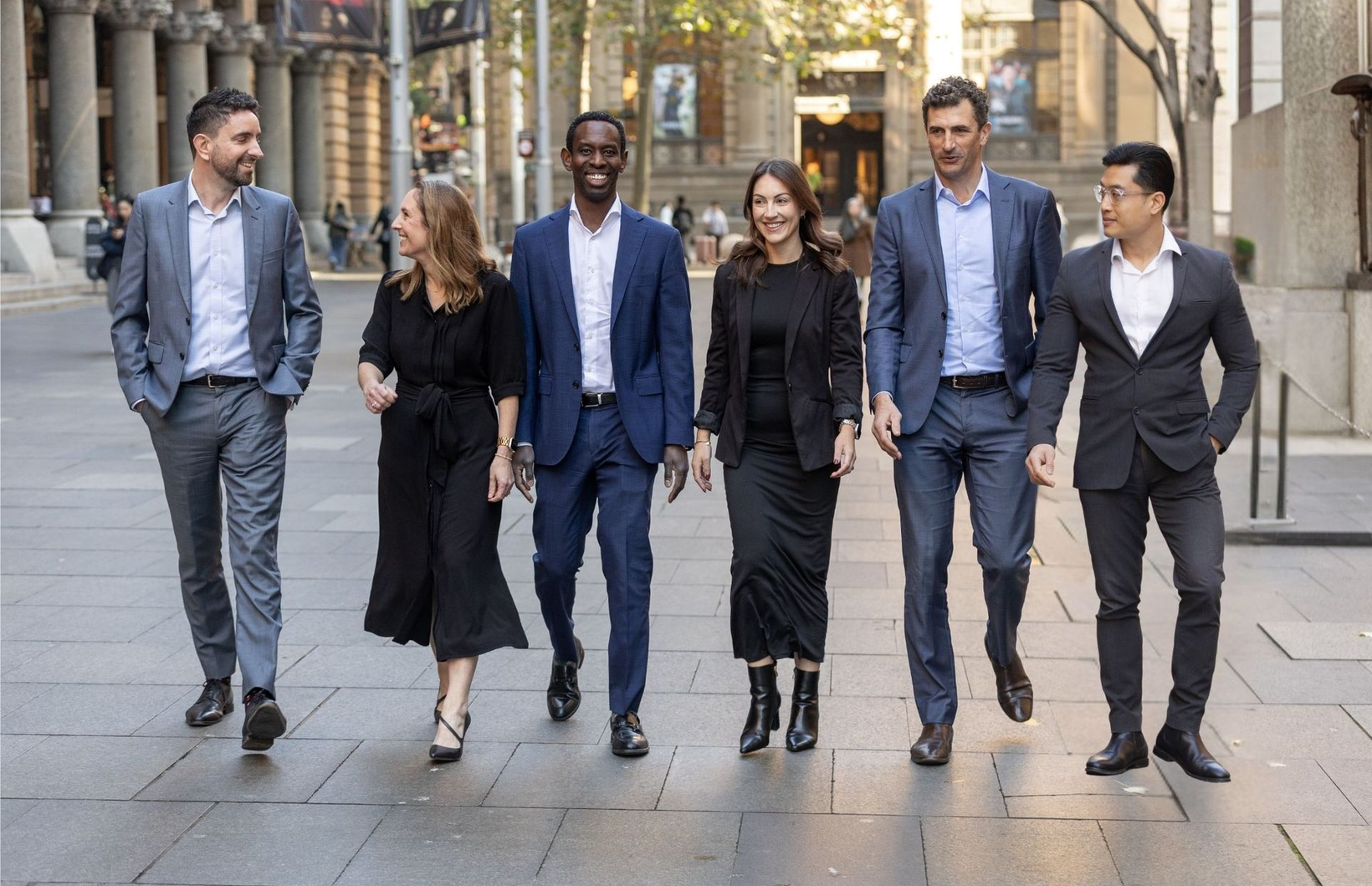 Six professionals walking together on a city sidewalk in business attire, smiling and chatting.
