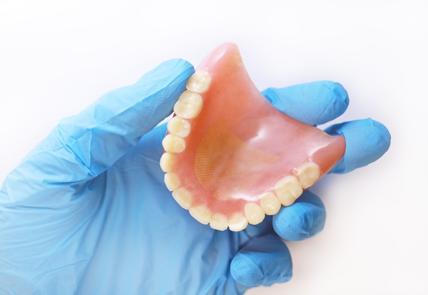 A Person Wearing Blue Gloves is Holding a Denture in Their Hand — Jaw Gear Dental Prosthetics in Moss Vale, NSW