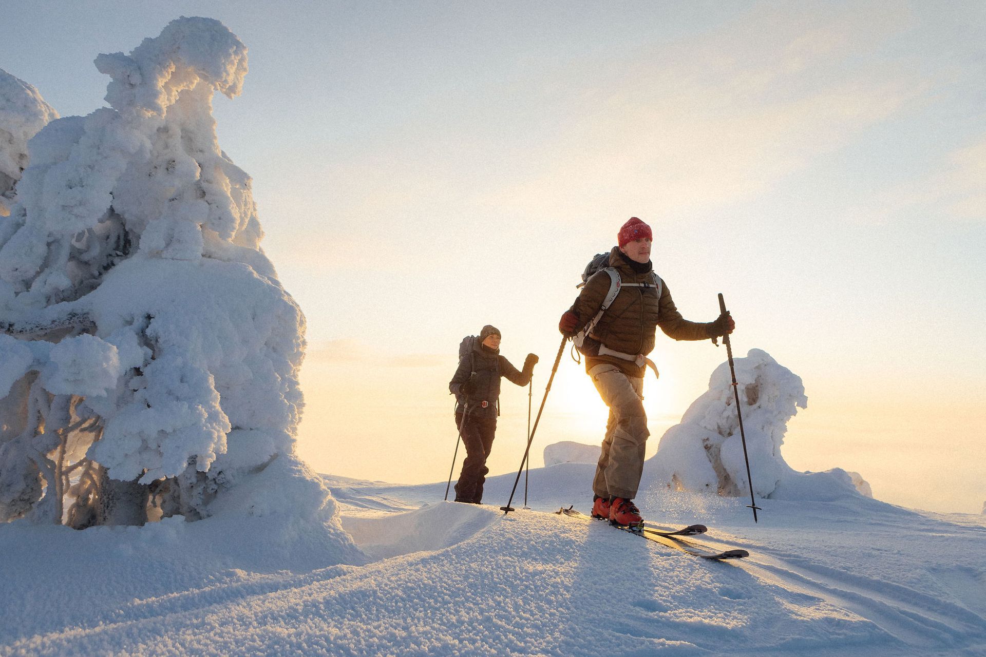 Kaksi skinnaajaa ylittää lumista harjannetta auringonnousussa, huurteisten puiden ja pitkien varjojen keskellä.