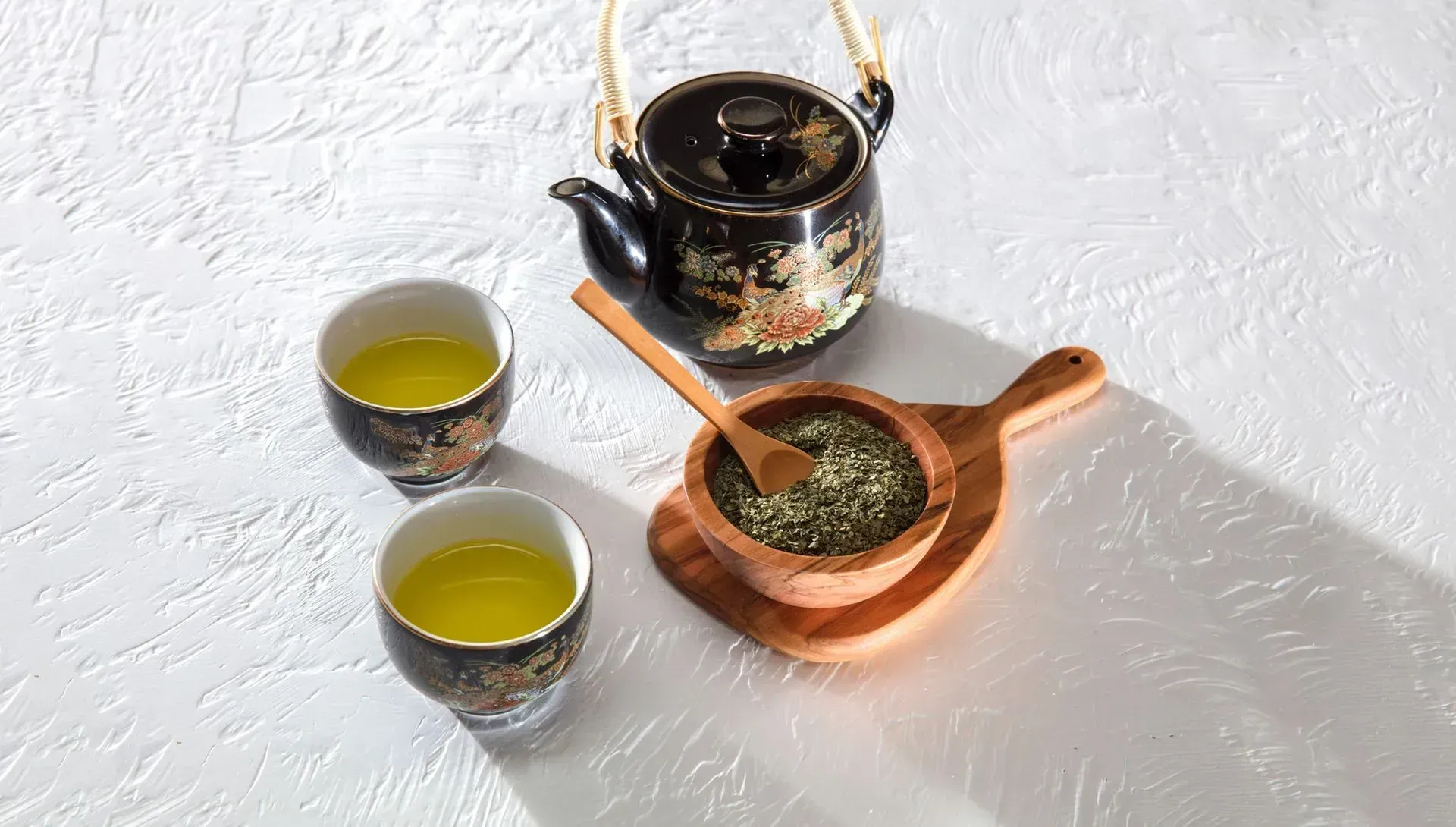 Tea set with two cups, a teapot, and a wooden scoop of loose tea on a white textured surface