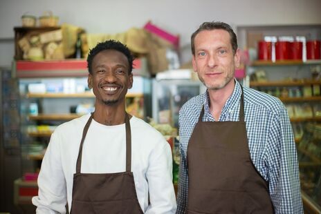 Two smiling coworkers in aprons standing behind a shop counter with shelves of goods behind them