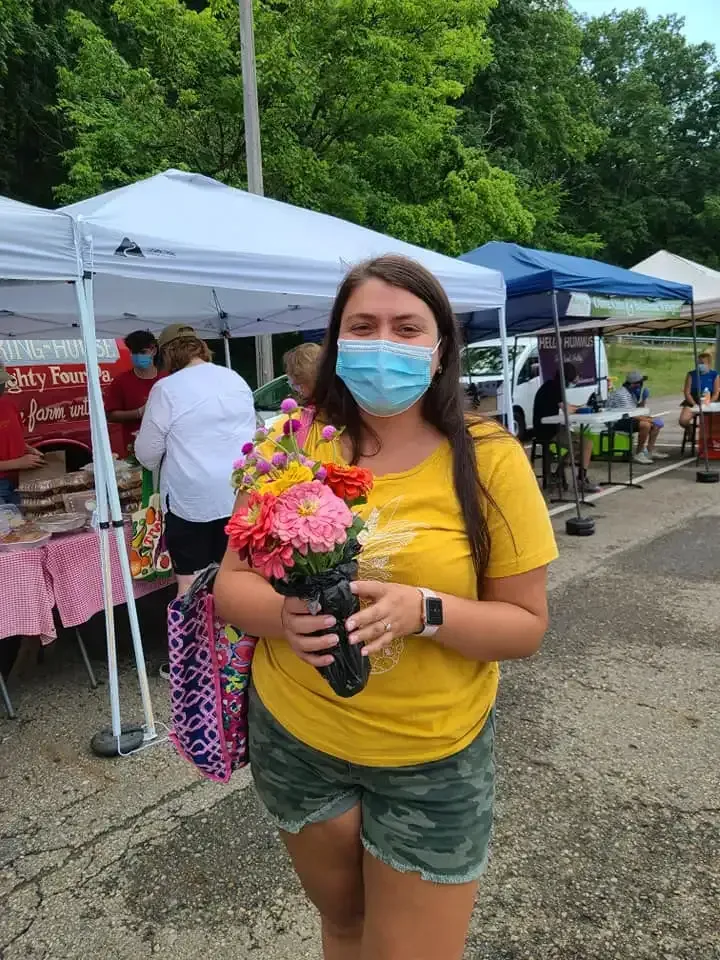 Woman wearing a mask holds a bouquet of flowers at an outdoor market with tents and vendors.