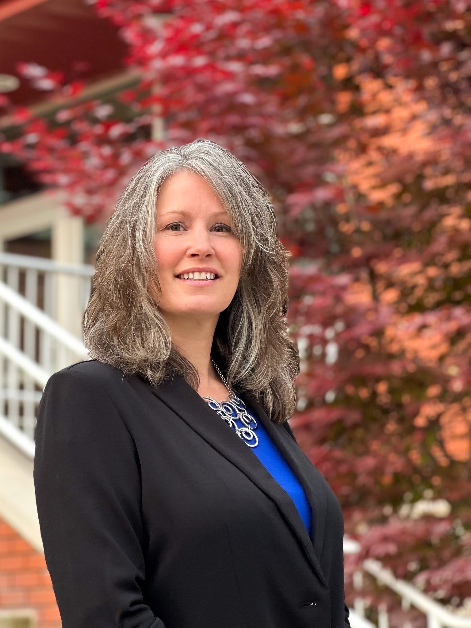 Woman in a black blazer and blue top standing outdoors in front of red autumn foliage and white stairs