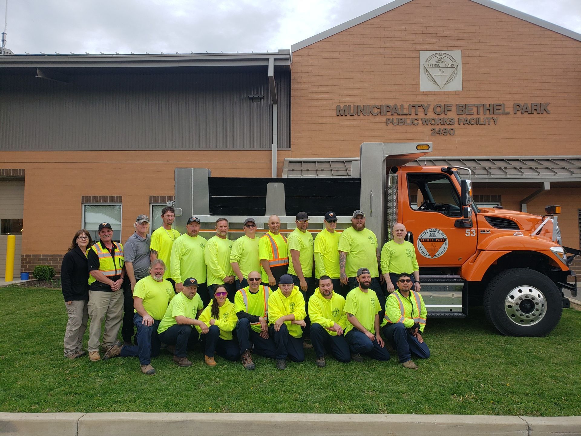 Group in neon yellow safety shirts posing beside an orange truck outside a municipal building