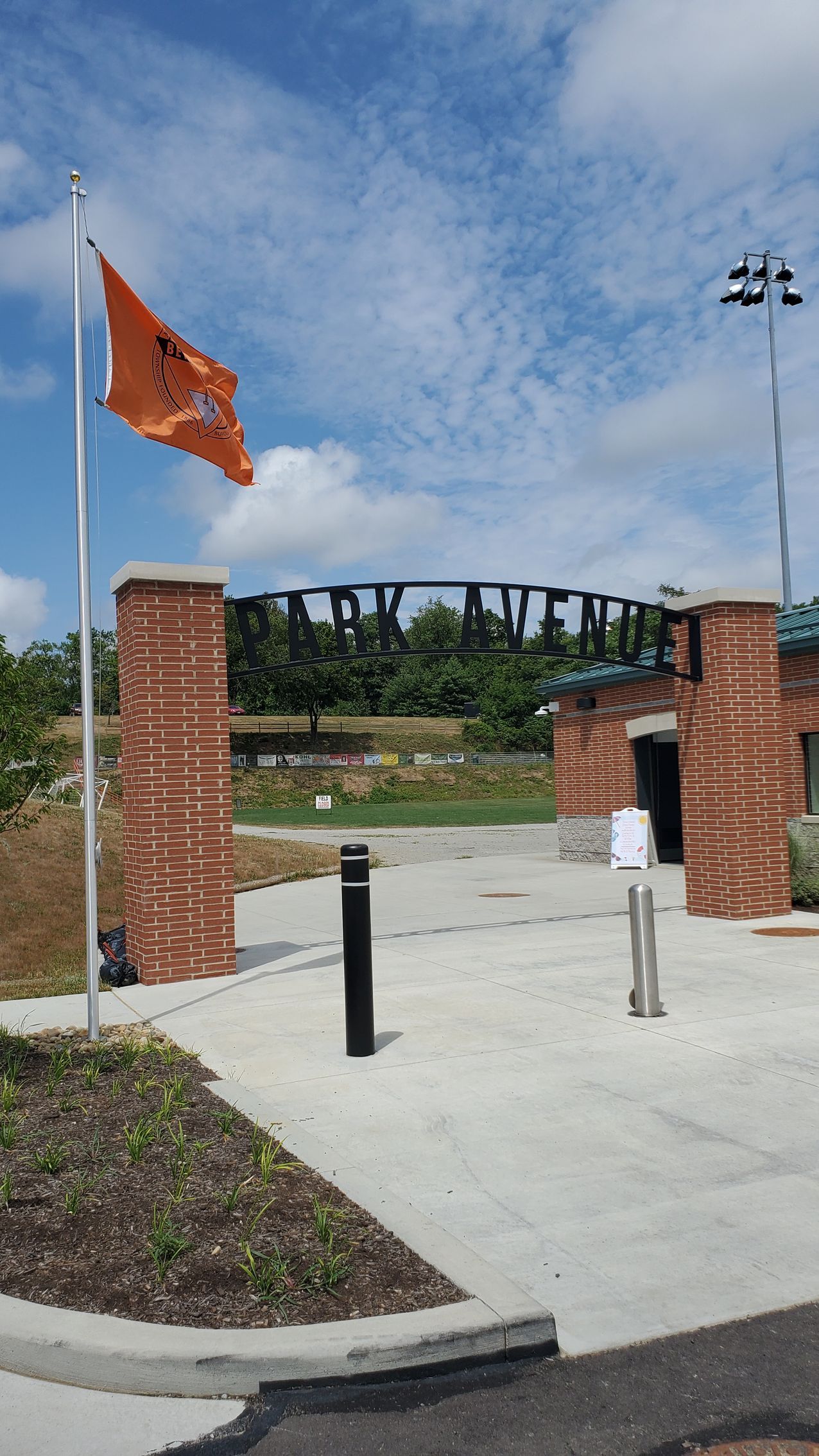 Brick entrance with orange flag, black bollards, and a path leading to a park bridge under a cloudy sky