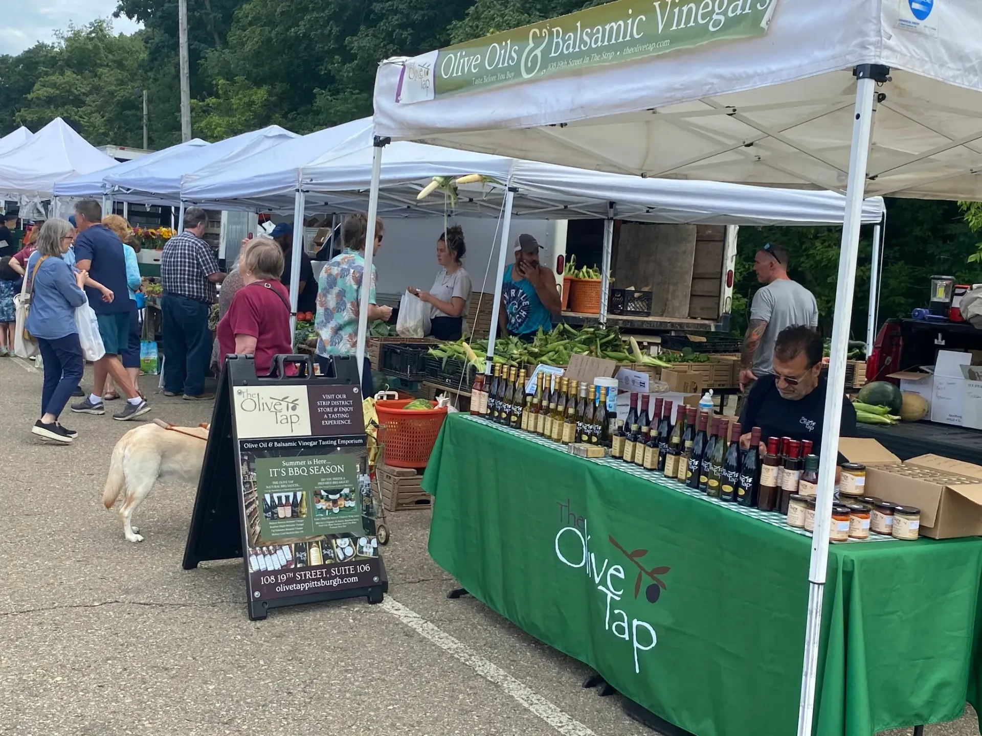 Outdoor market stall with green tablecloths, bottles, and a chalkboard sign under white tents