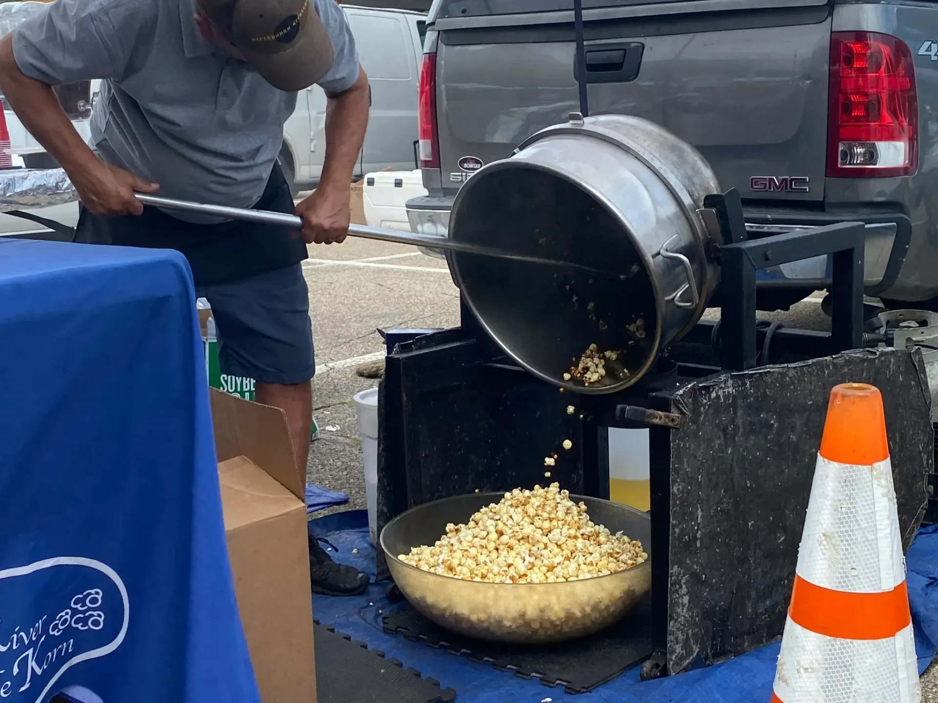 Person pours popcorn from a large metal drum into a bowl beside a black pickup truck at an outdoor event.