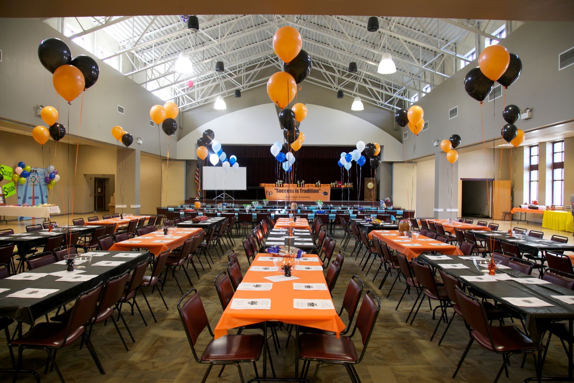 Banquet hall set with long tables, orange tablecloths, and black-orange balloons under high arched ceilings