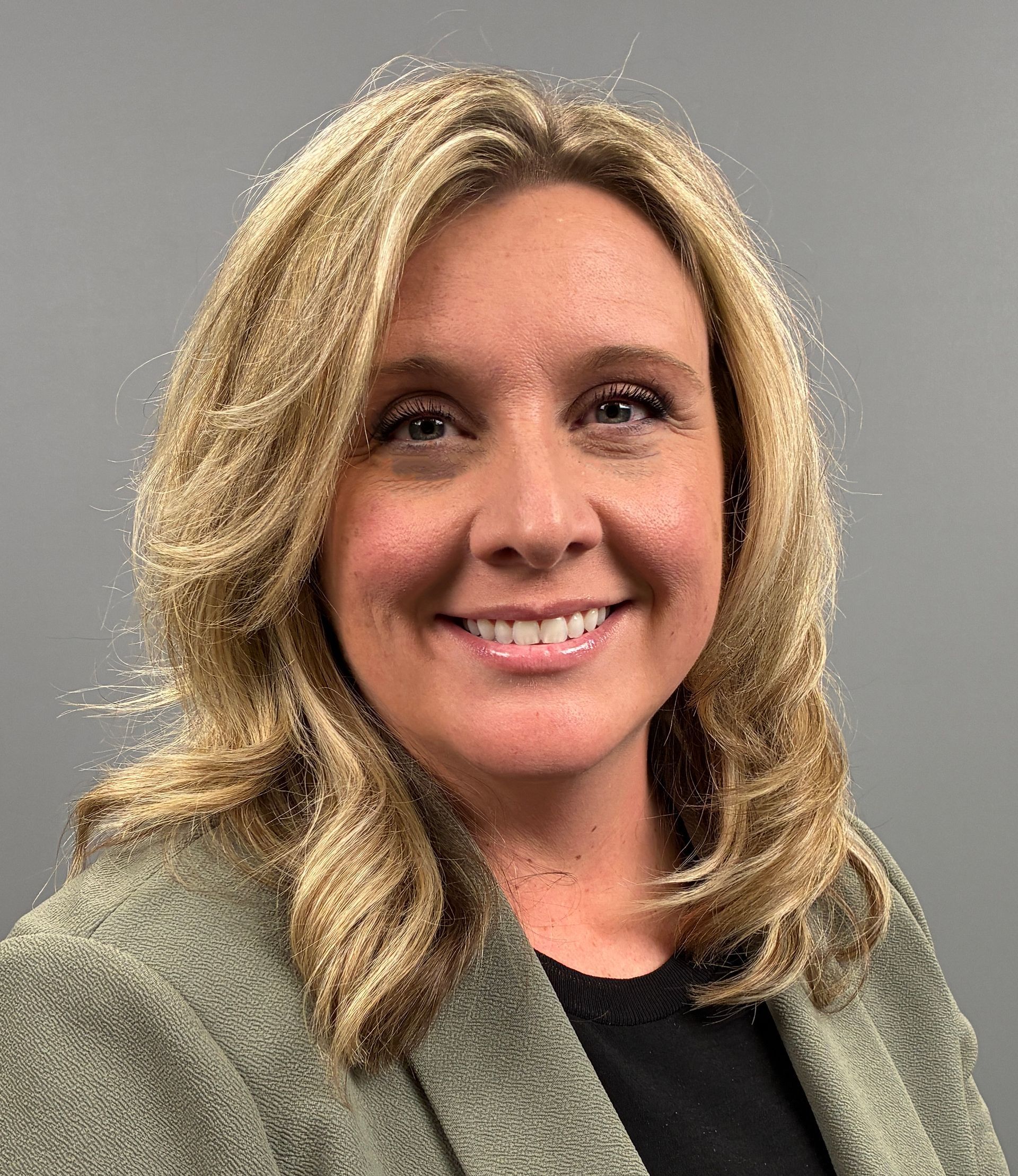 Smiling woman in a gray blazer with blonde hair against a plain gray background