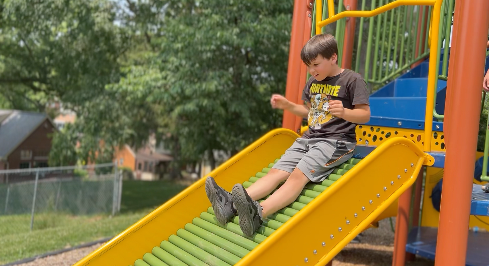 Boy seated on a yellow playground slide, looking at a device, with trees and a fence behind him