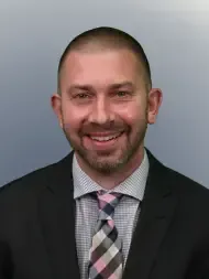 Smiling man in a dark suit, patterned shirt, and pink plaid tie against a gray background