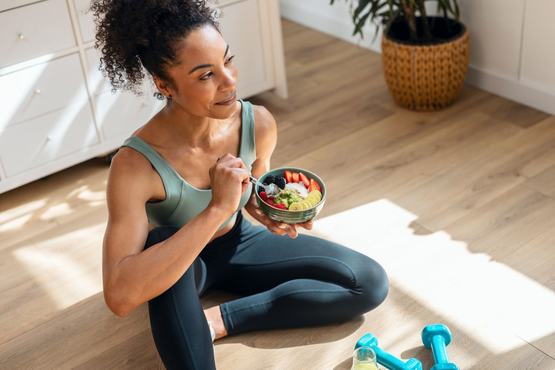 Woman sitting on a sunlit floor, eating a colorful salad next to blue dumbbells and a yoga mat.