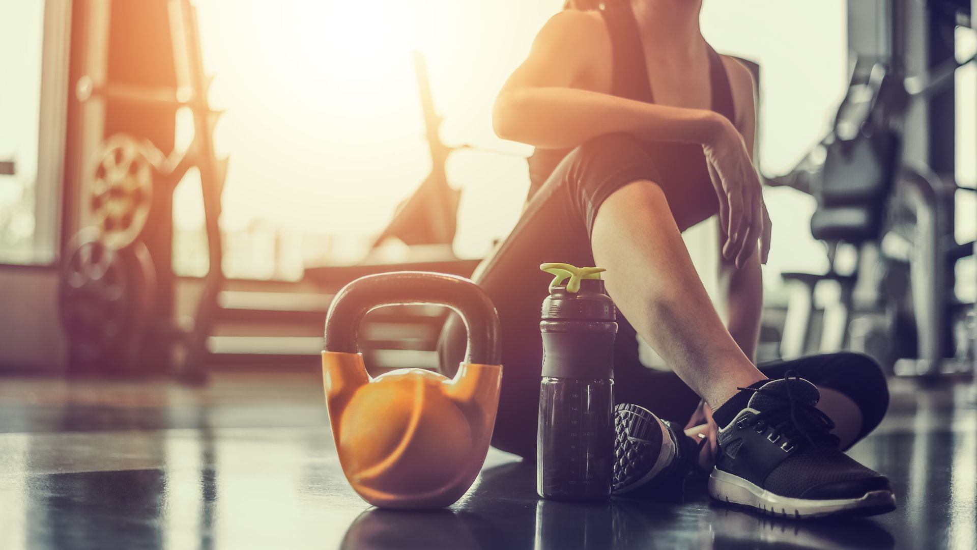 Gym floor with kettlebell, water bottle, and a person tying shoelaces in warm sunlight