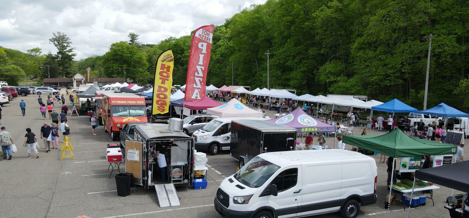 Outdoor market with vendor tents, banners, and parked vans along a tree-lined road