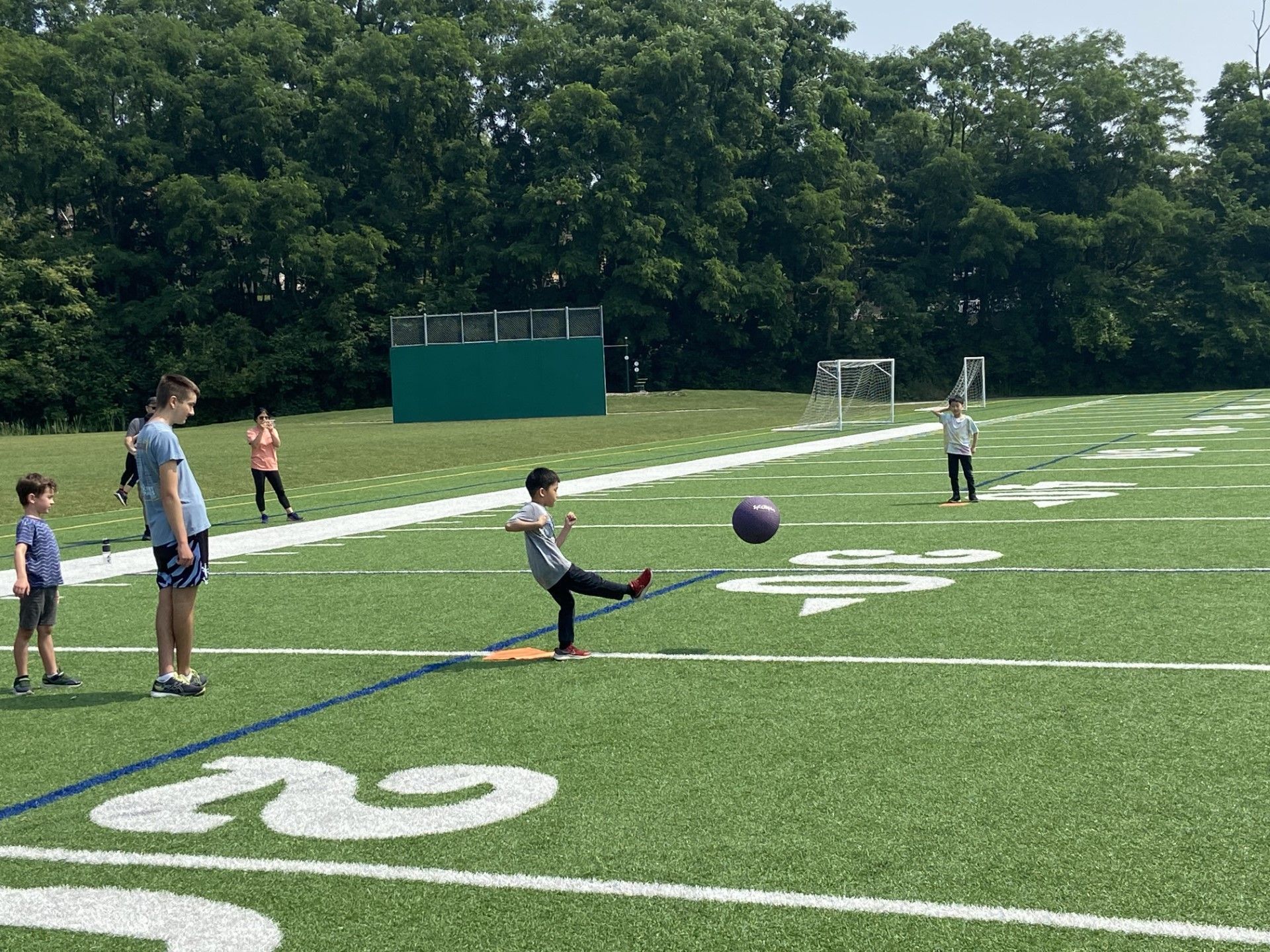 Children playing kickball on a grassy field with white yard lines and trees in the background