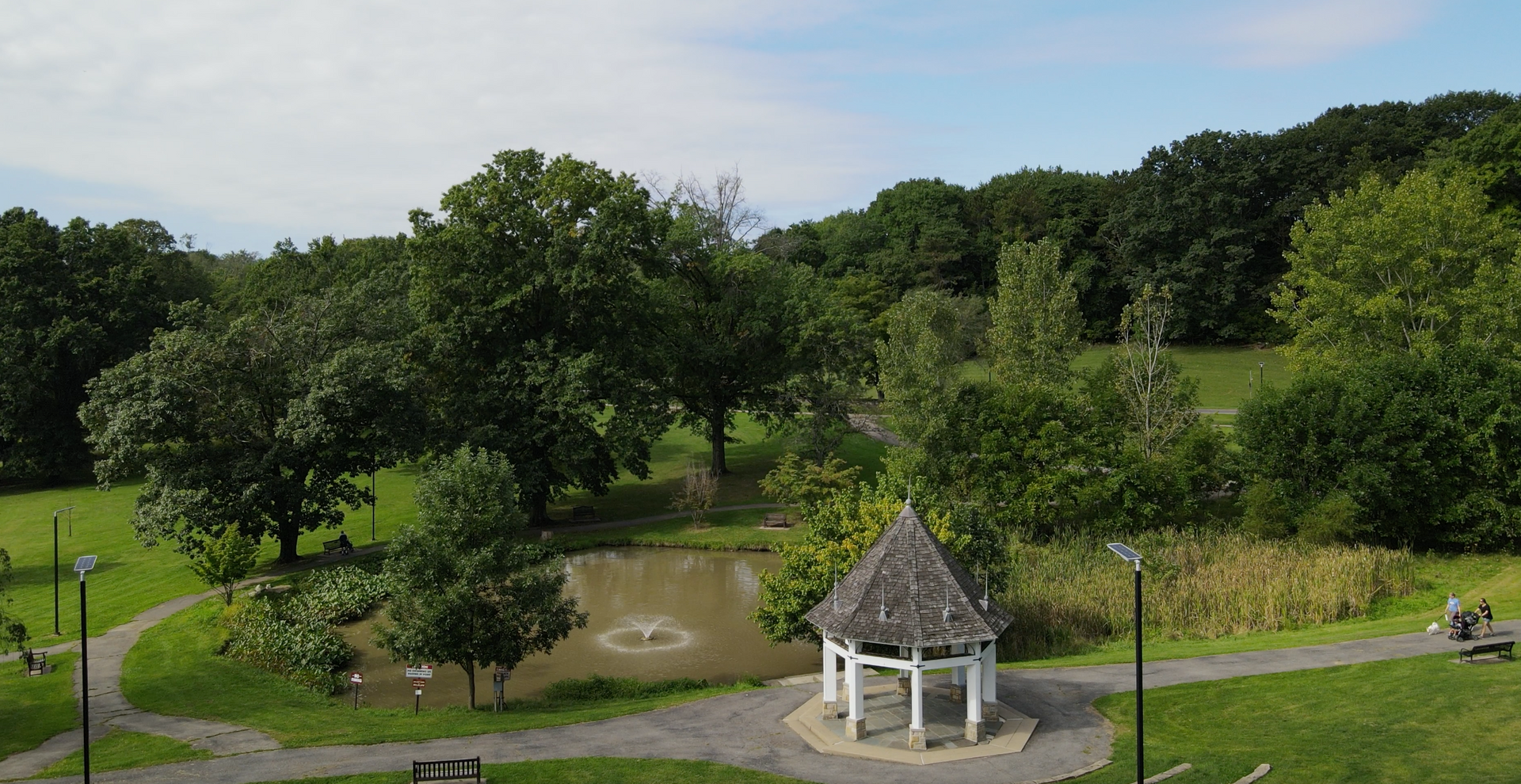 Green park with trees, a small pond, and a white gazebo on a grassy hillside
