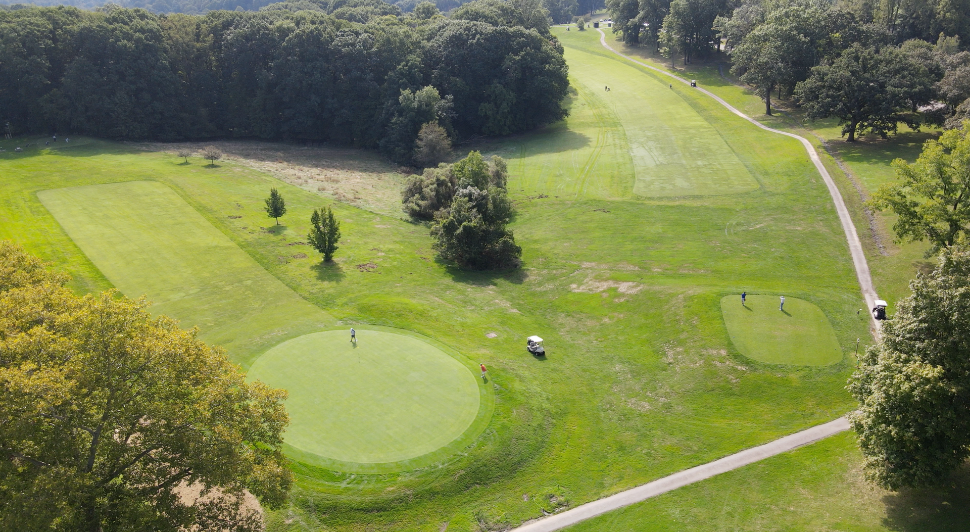Aerial view of a green golf course with fairways, trees, paths, and a sand bunker