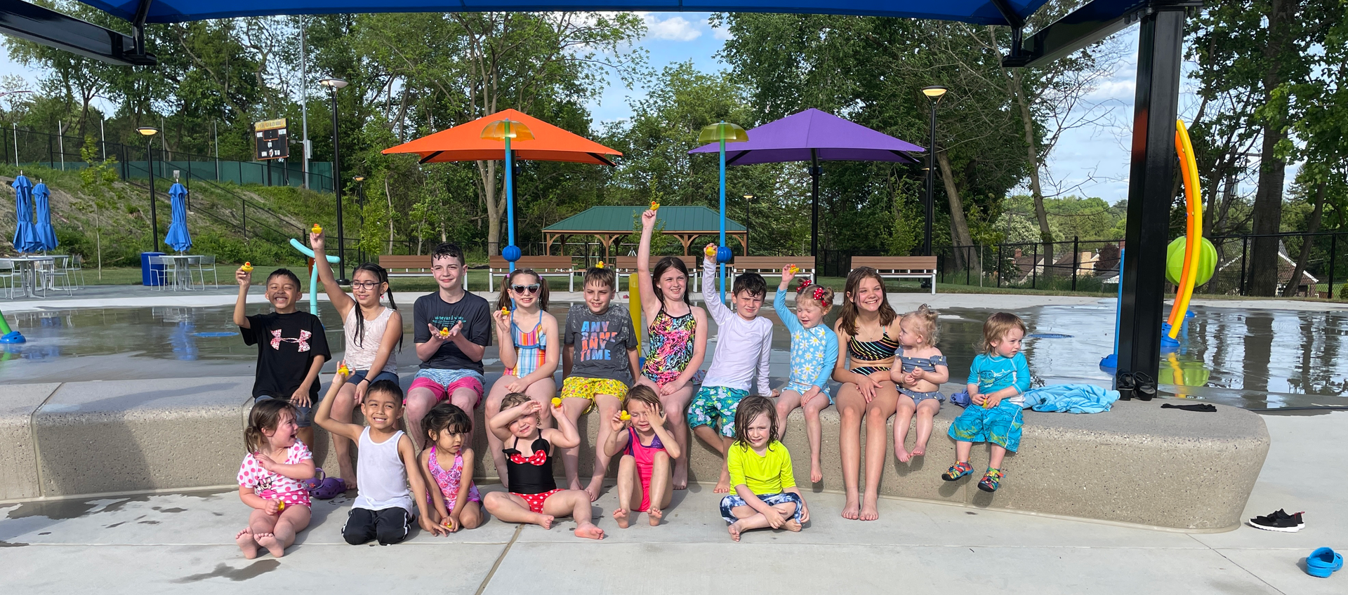 Group of children posing by a pool with colorful umbrellas and trees in the background.