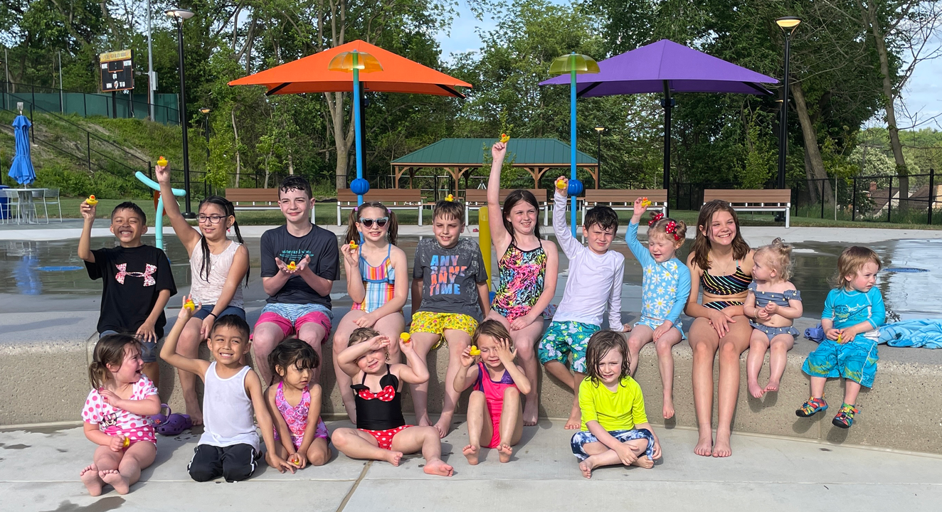 Group of children posing together outdoors in swimsuits under colorful umbrellas by a pool.