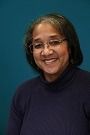 Smiling person in glasses and a dark blue turtleneck against a plain dark background