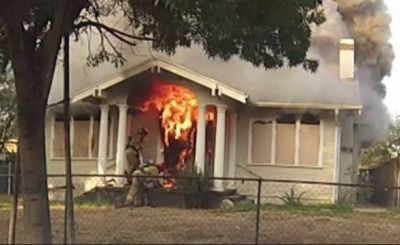 House fire with flames bursting from the front doorway of a white home, smoke rising above.