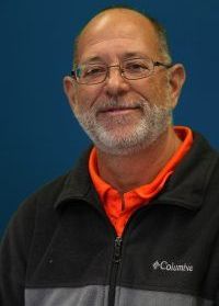 Smiling man with glasses wearing a black jacket and orange shirt against a blue background