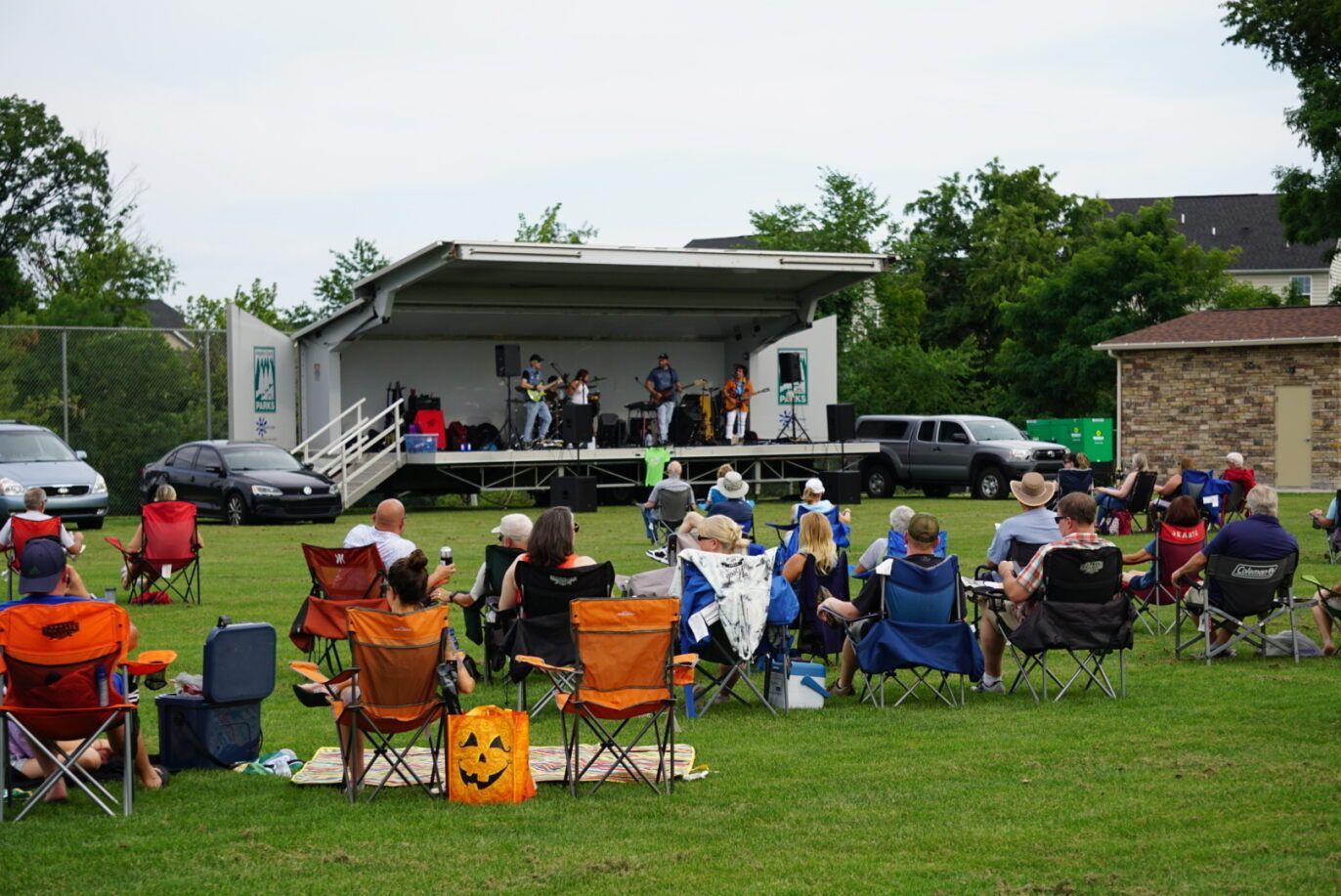 Outdoor crowd seated in folding chairs watching a live performance on a small stage in a park