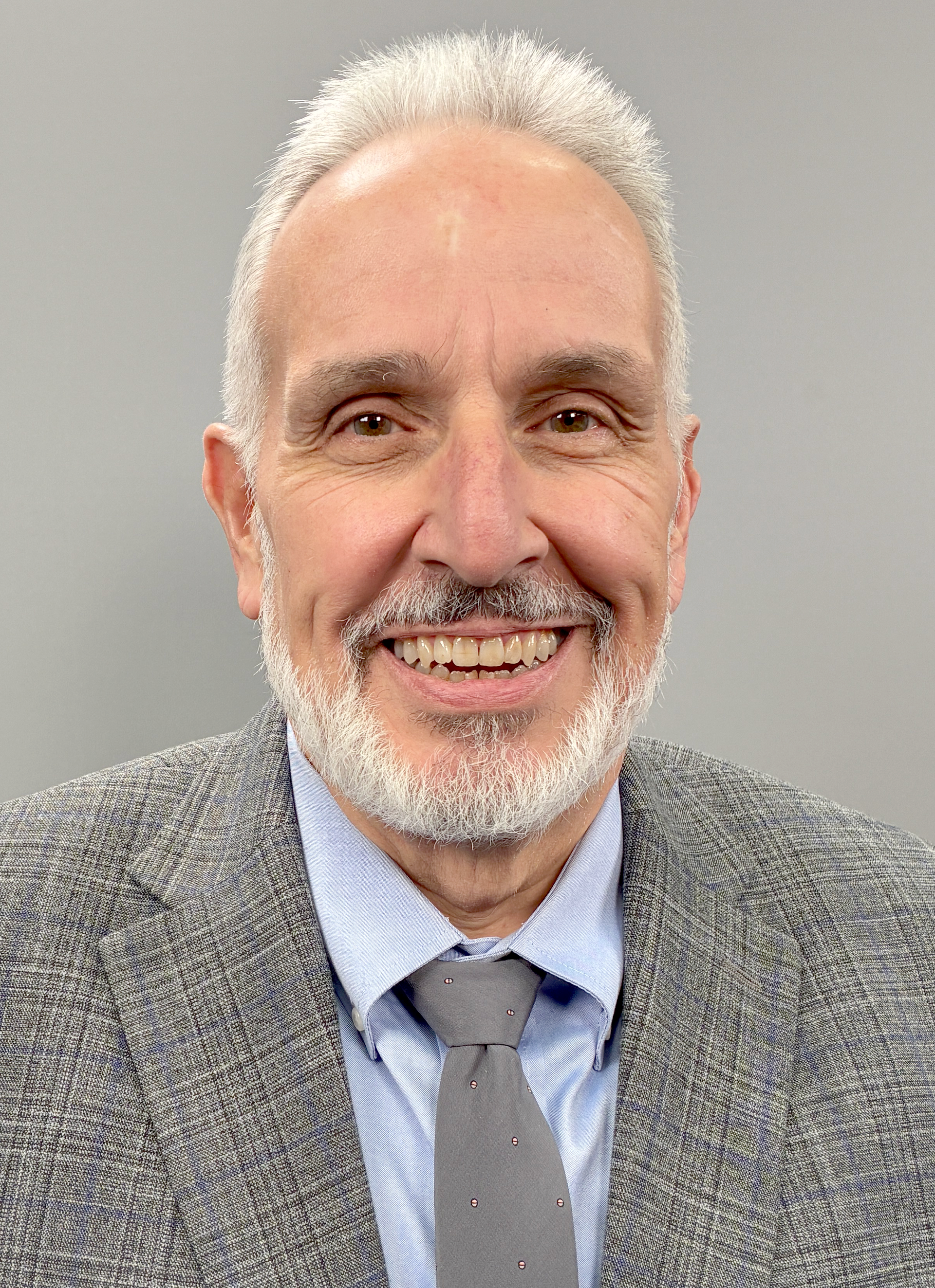Smiling man in a gray blazer, blue shirt, and tie against a plain gray background