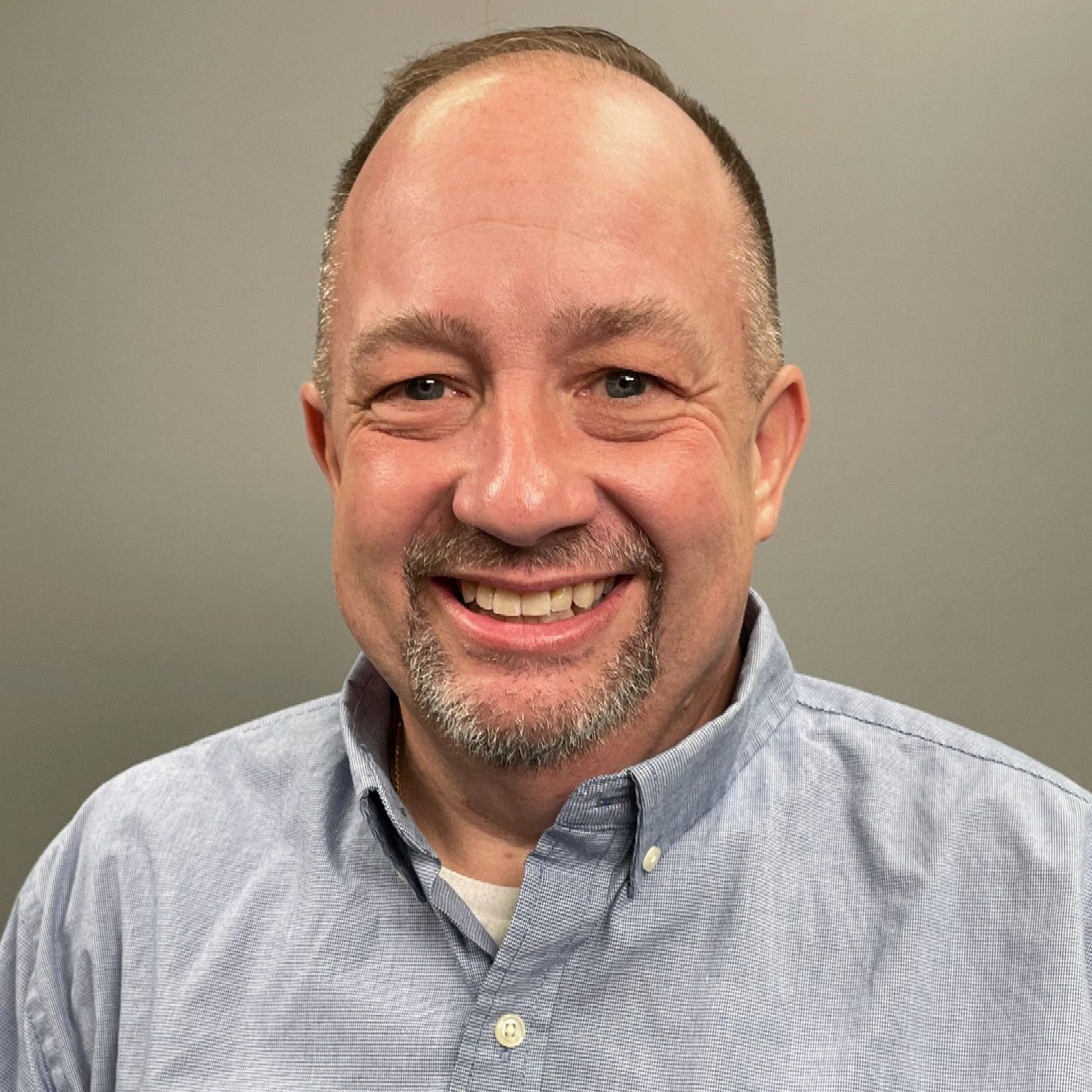 Smiling man in a light blue button-down shirt against a plain gray background