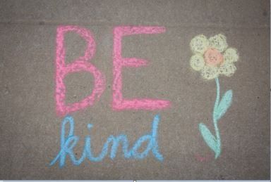 Chalk message “BE kind” in pink and blue with a small flower drawing on dark pavement