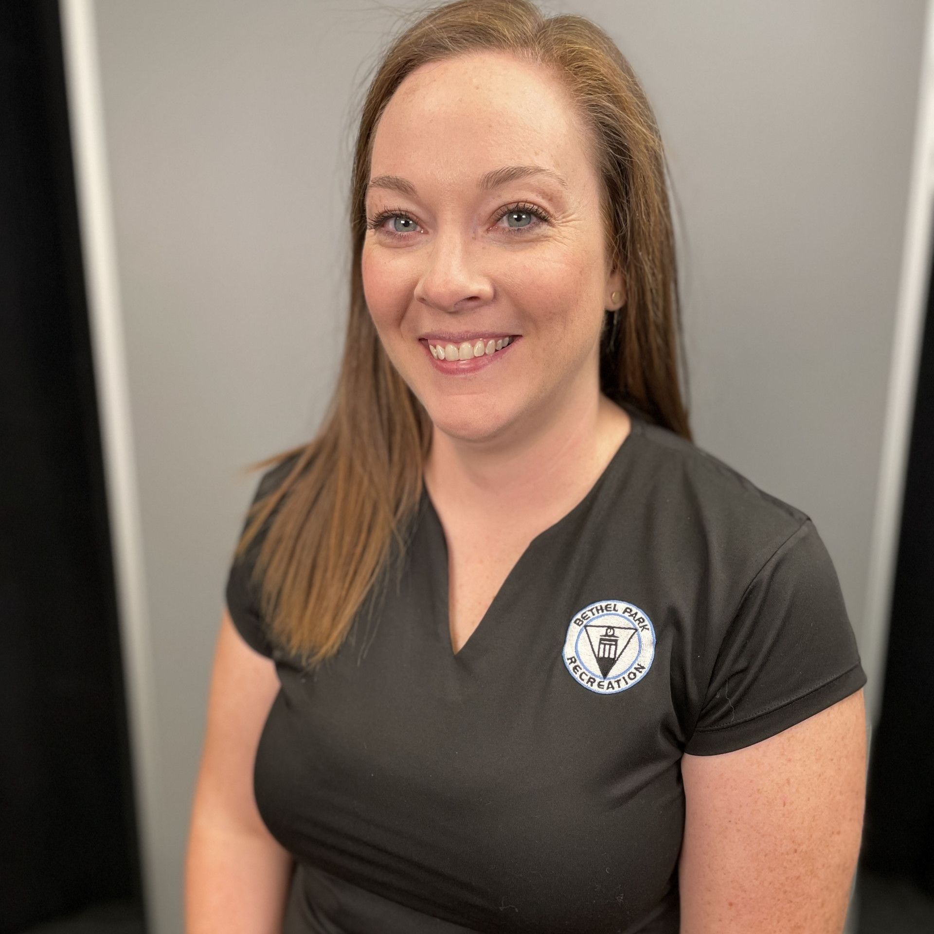 Smiling woman in a black shirt with a circular logo, standing indoors against a gray background.