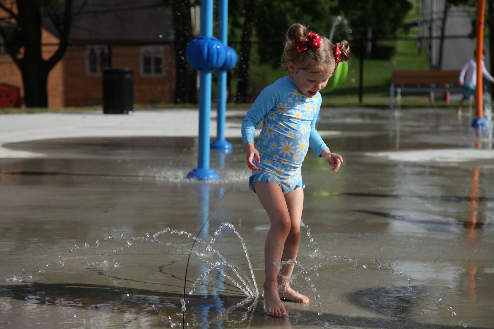 Child in blue swimsuit splashing in a wet park splash pad on a sunny day